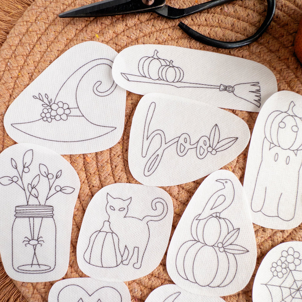 White water-soluble Halloween stick and stitch embroidery patterns, like a witch hat, ghost, and cat, on a woven mat.