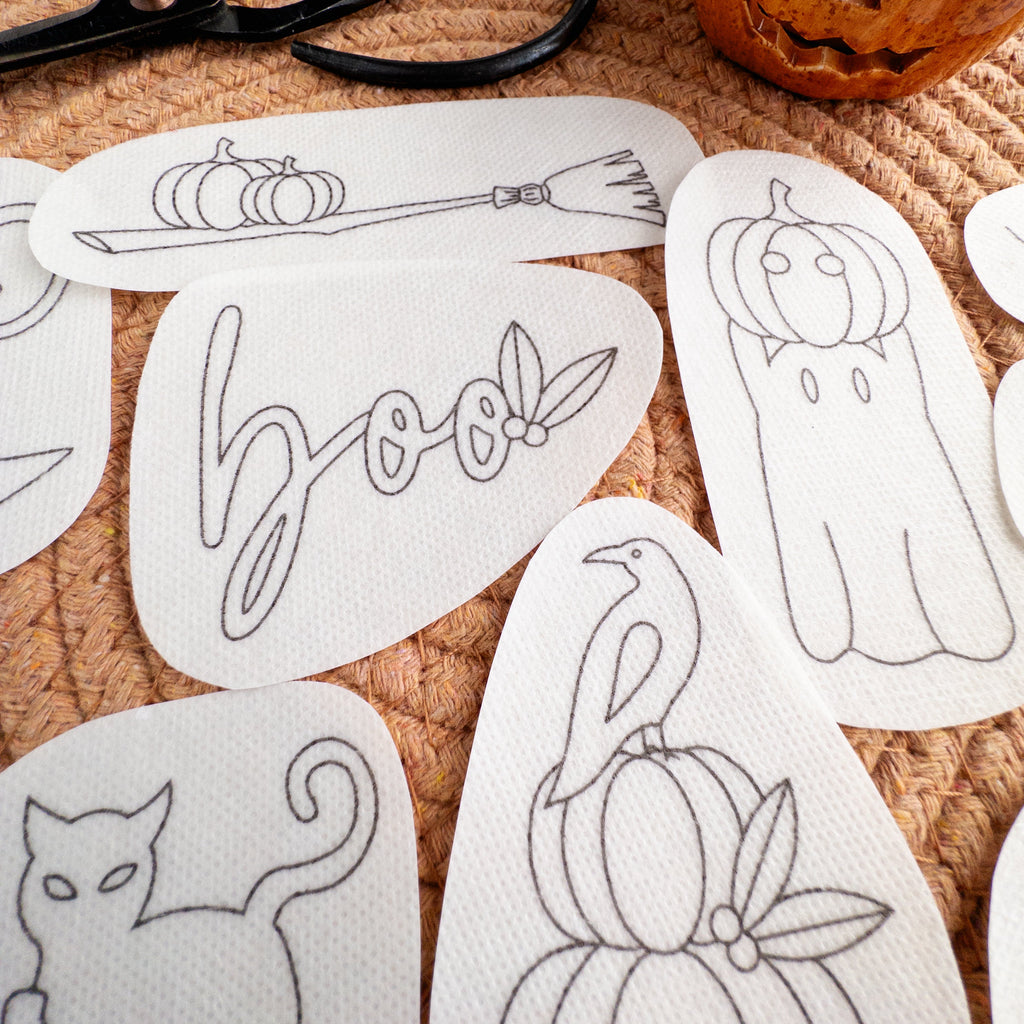 White water-soluble embroidery patterns: ghost, pumpkin, cat, broom, raven, and "boo" on a woven mat.