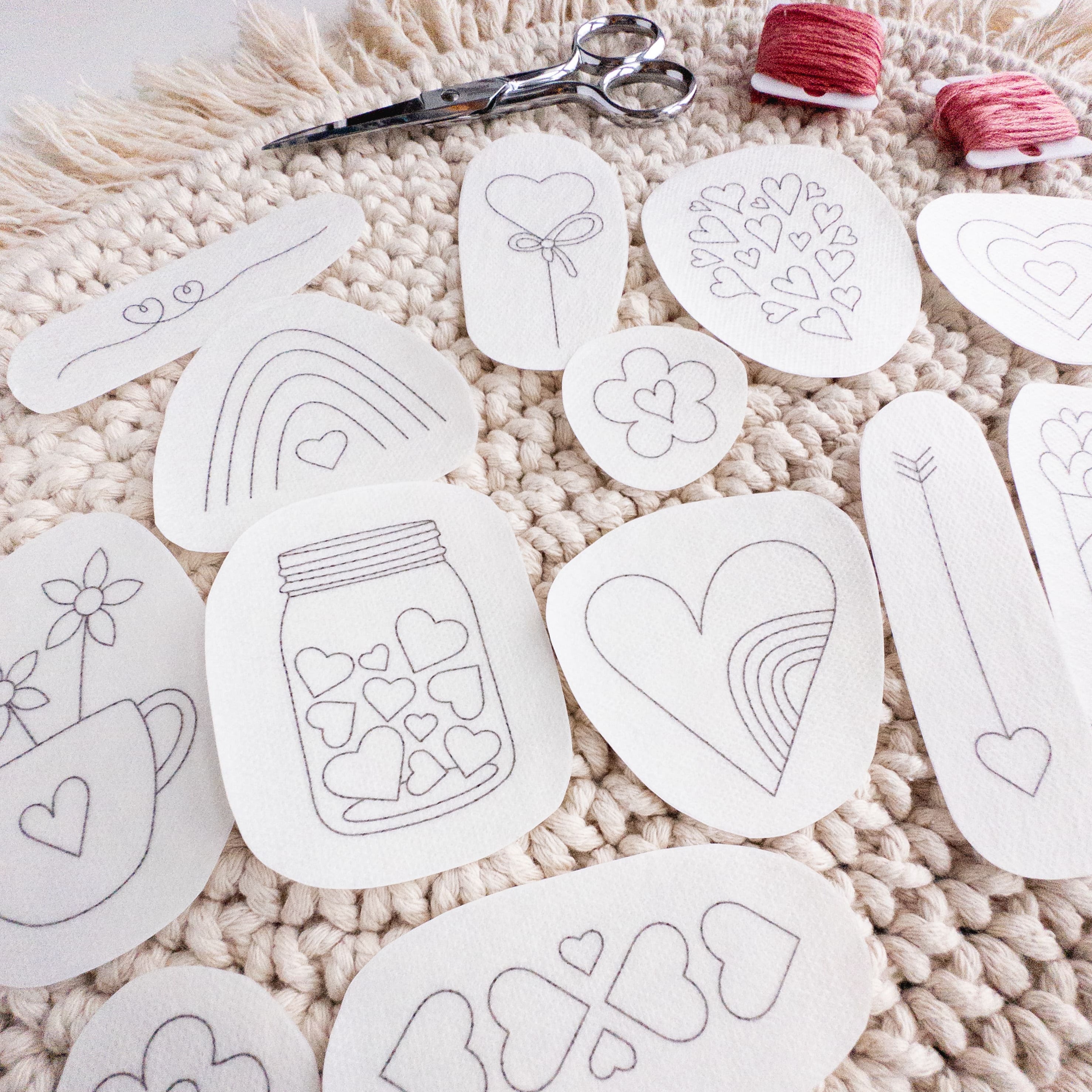 13 white water-soluble heart embroidery patterns, various sizes, on a cream crocheted mat with scissors and red floss.