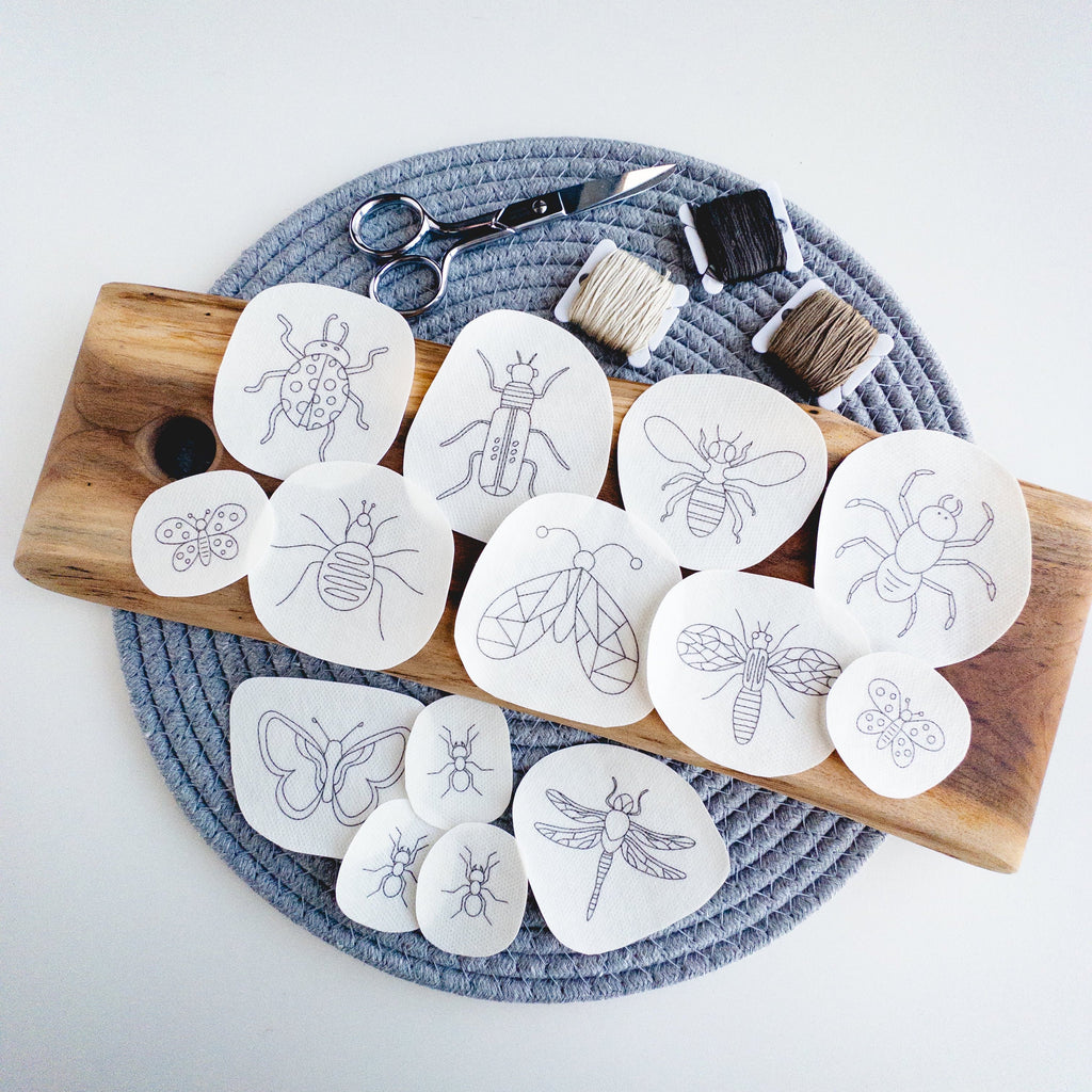 14 white water-soluble insect embroidery patterns on a wooden board with thread and scissors.