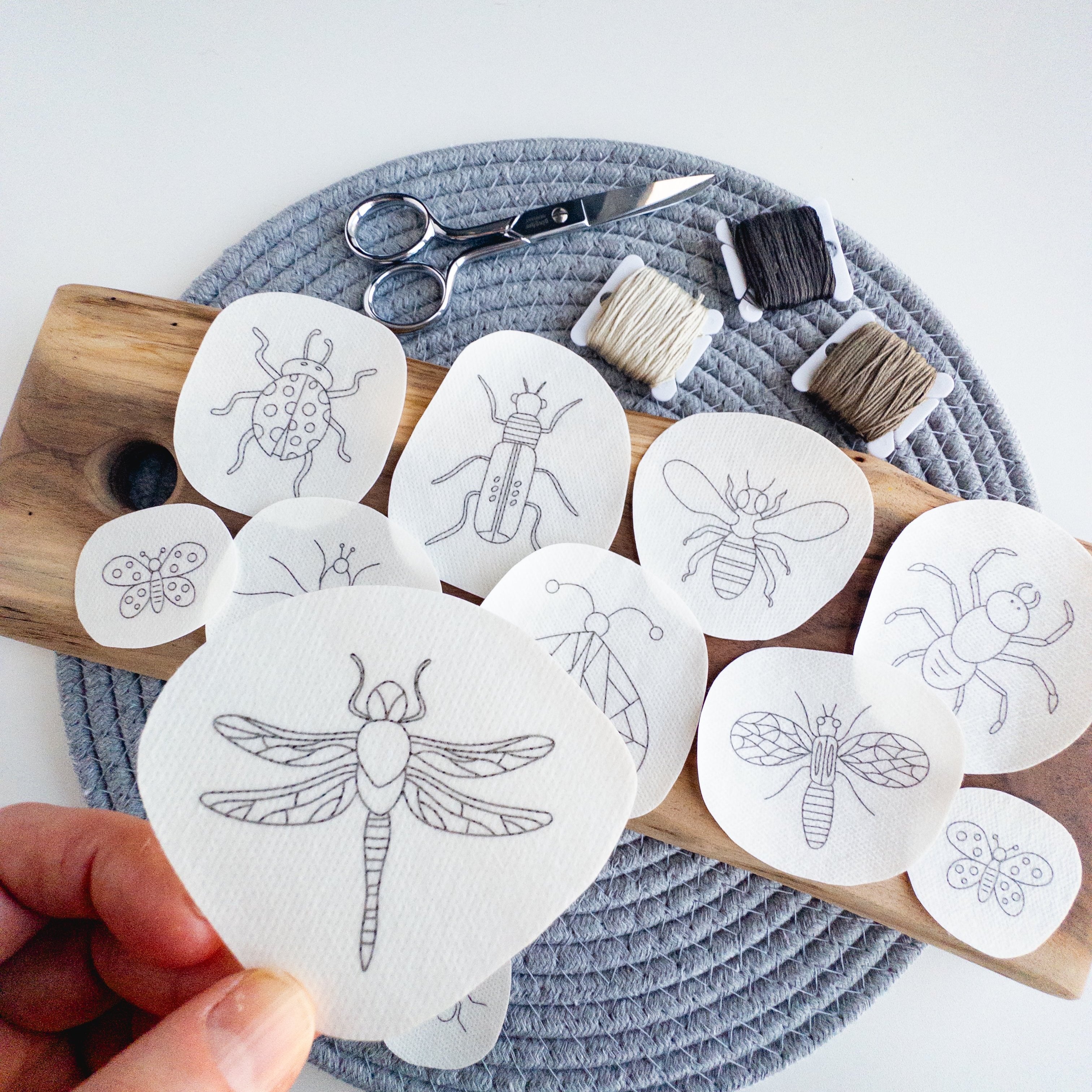 Assorted insect embroidery patterns on white water-soluble stabilizer patches with scissors and floss.