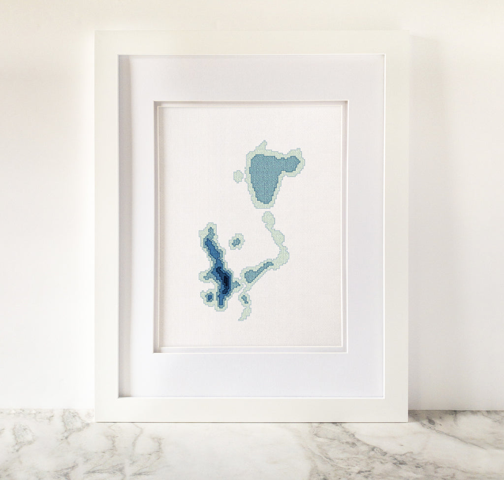 Framed Okoboji Iowa Lakes cross-stitch pattern in shades of blue on white fabric.