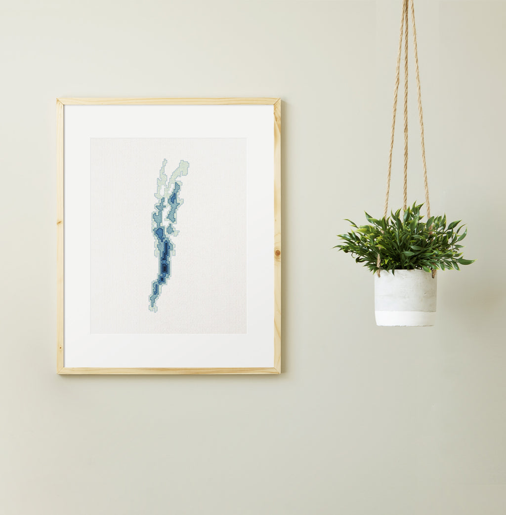 Framed Lake Champlain cross-stitch pattern in shades of blue and green, displayed on a light wall next to a hanging plant.