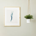 Framed Lake Champlain cross-stitch pattern in shades of blue and green, displayed on a light wall next to a hanging plant.