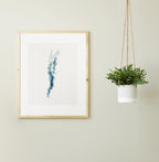 Framed Lake Champlain cross-stitch pattern in shades of blue and green, displayed on a light wall next to a hanging plant.
