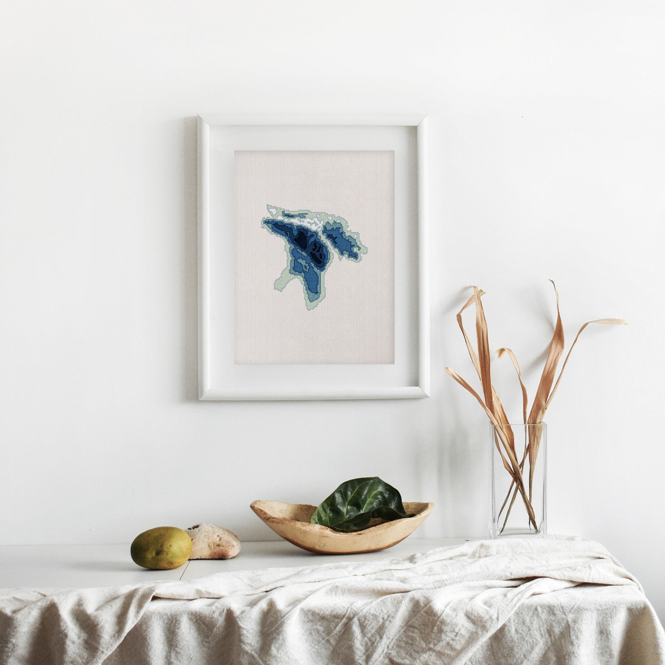 Framed Lake Huron cross-stitch pattern in blue and white, displayed on a minimalist white wall.