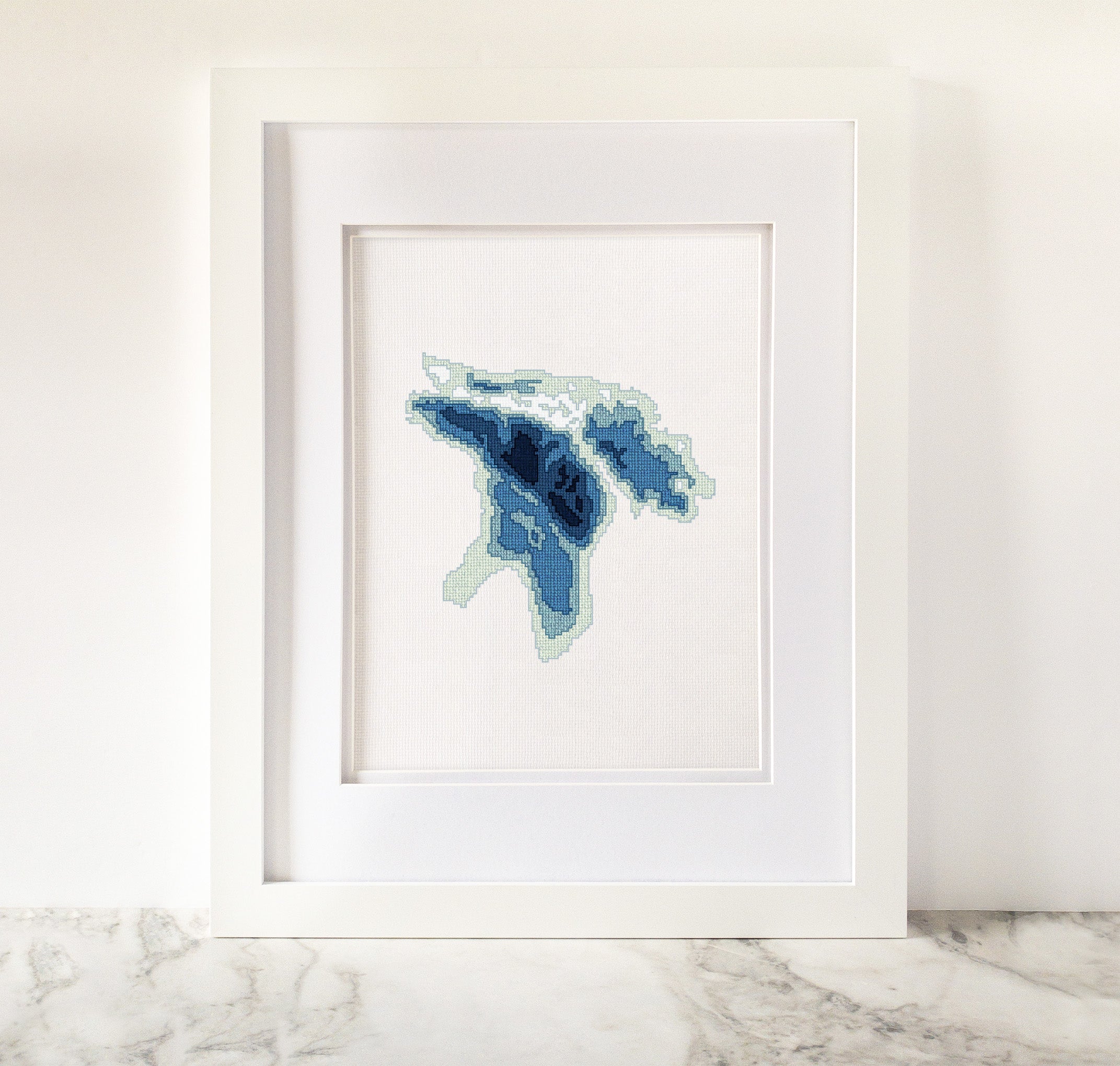 Framed Lake Huron cross-stitch art in shades of blue and green on white fabric.
