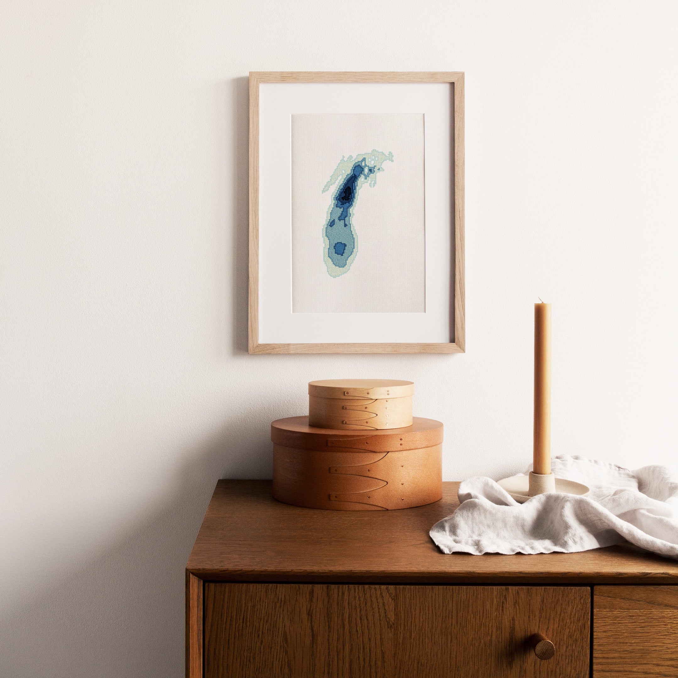 Framed Lake Michigan cross-stitch pattern in shades of blue, displayed above wooden boxes and a candle.