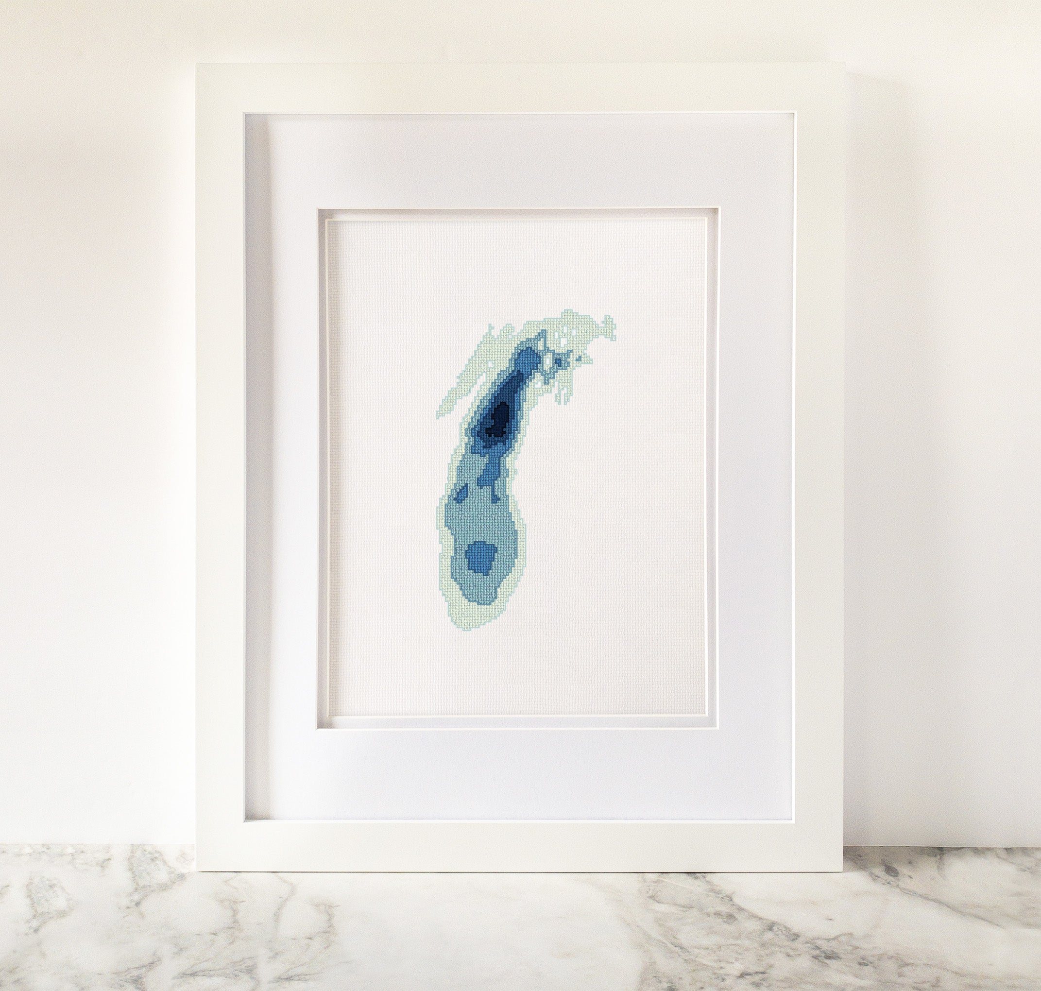 Framed Lake Michigan cross-stitch art with blue bathymetric map design on white fabric.