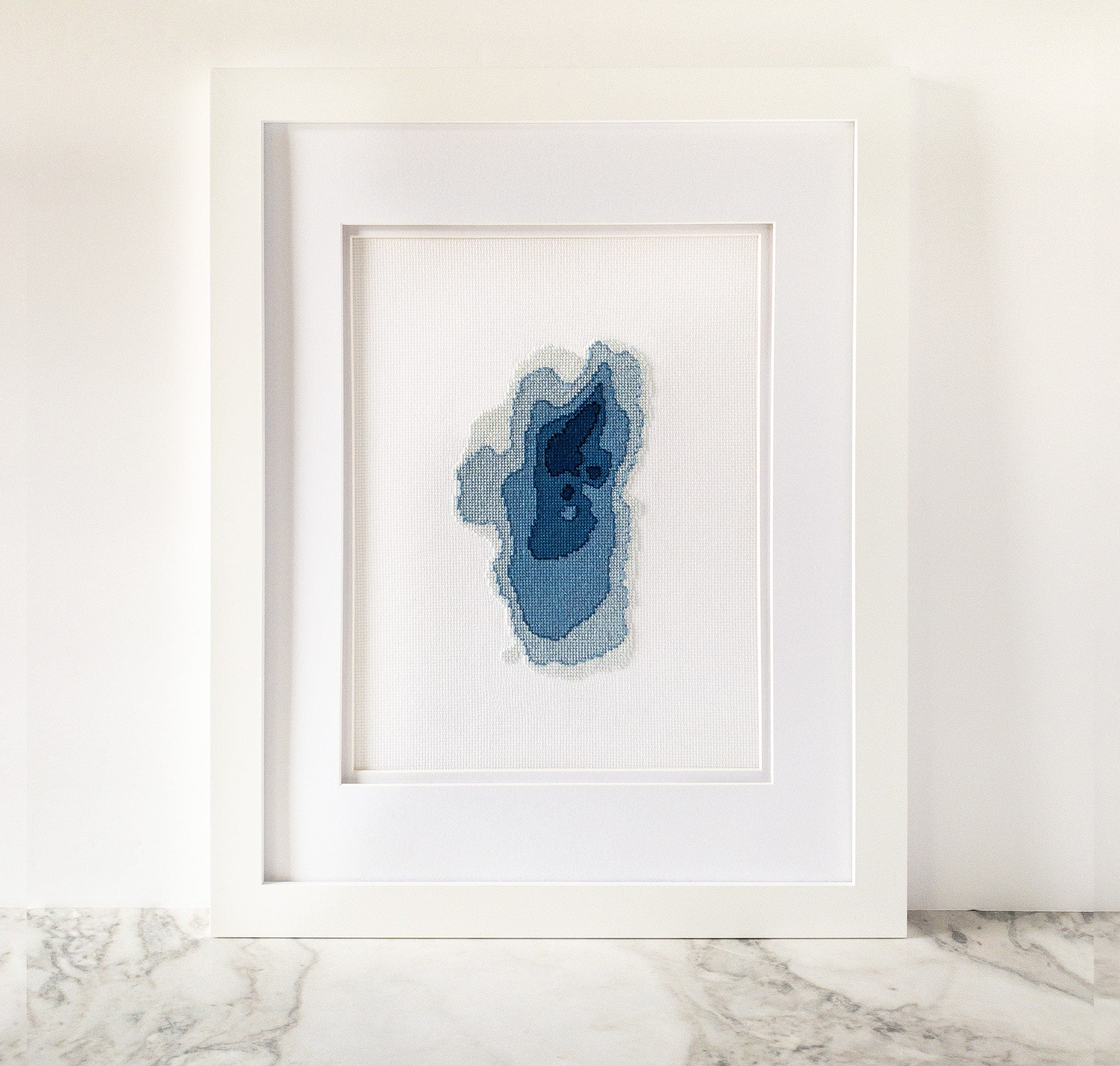 Framed Lake Tahoe cross-stitch art with blue bathymetric map design on white fabric.