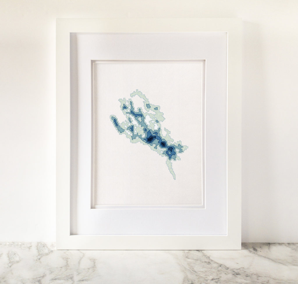 Framed Lake Winnipesaukee cross-stitch art with minimalist blue bathymetric map on white fabric.