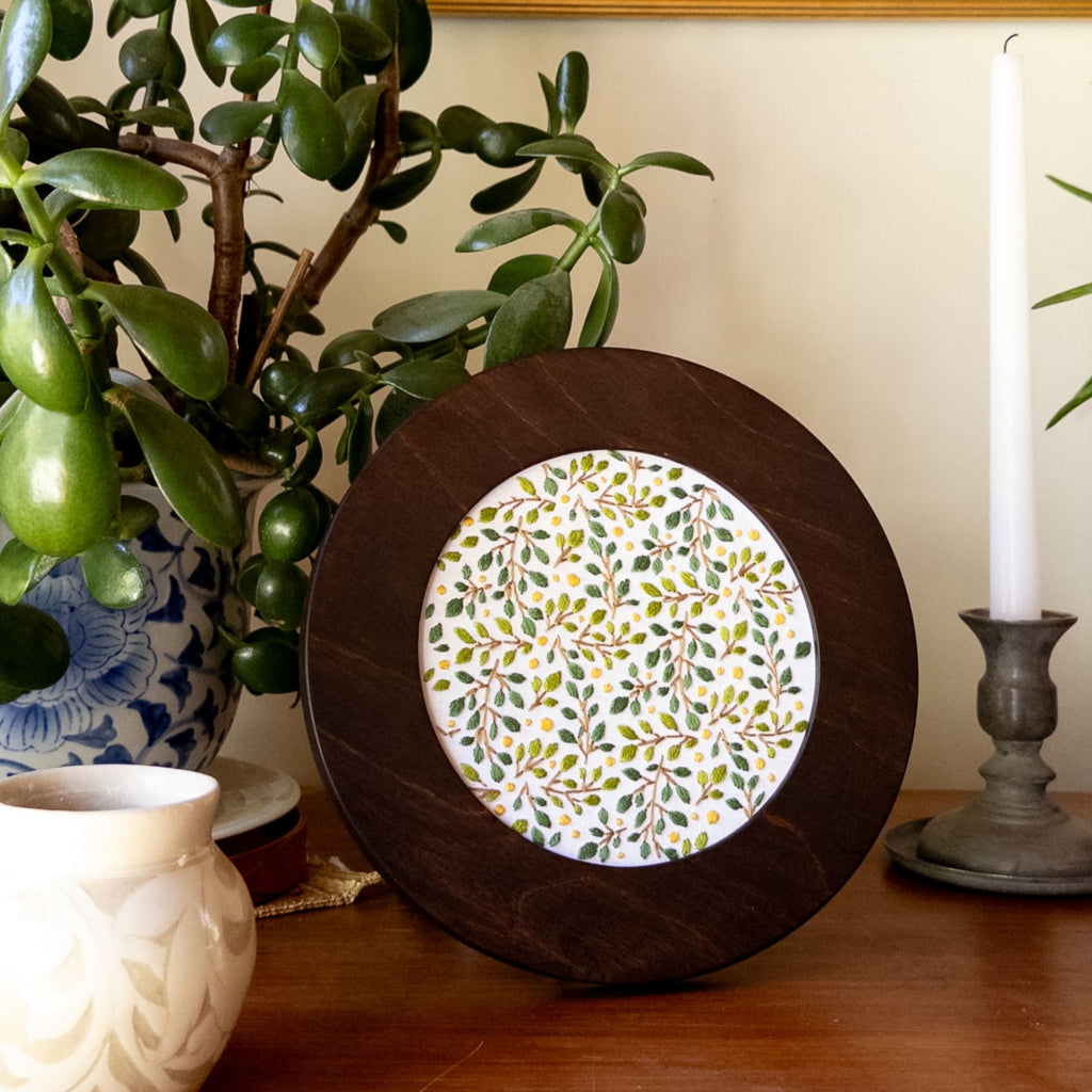 Round dark wood frame with a white embroidered lemon grove pattern of green leaves and yellow dots.
