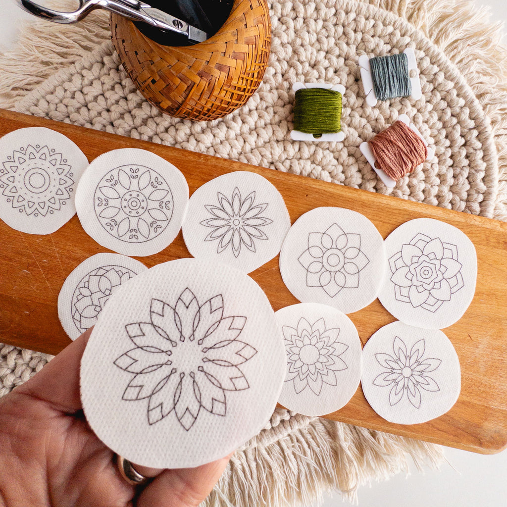Hand holding a round, white stick and stitch mandala pattern, with 11 more on a wooden board.