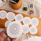 Hand holding a round, white stick and stitch mandala pattern, with 11 more on a wooden board.