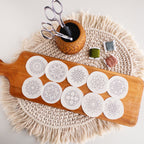 12 white circular mandala stick and stitch embroidery patterns on a wooden board with scissors and floss.