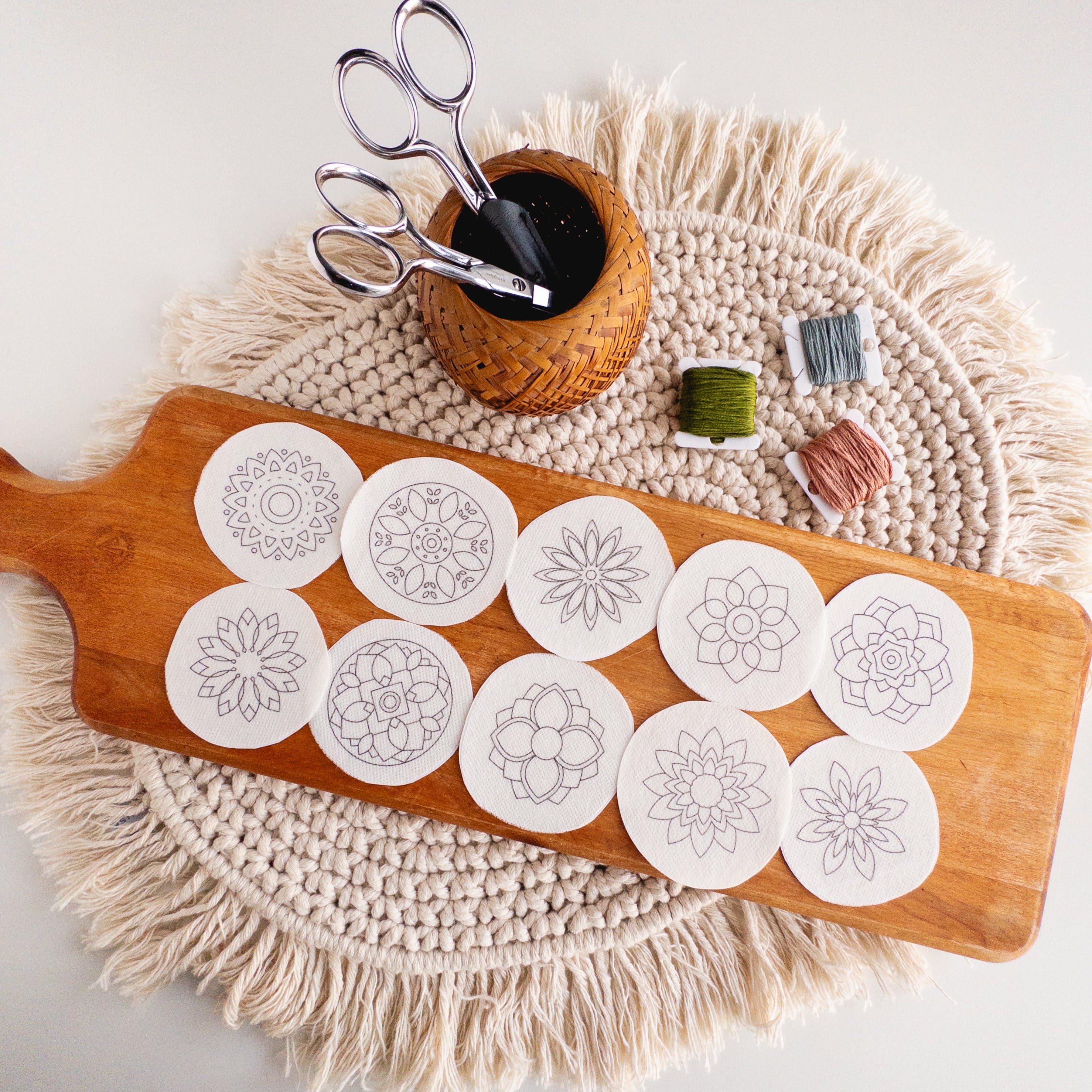 12 white circular mandala stick and stitch embroidery patterns on a wooden board with scissors and floss.
