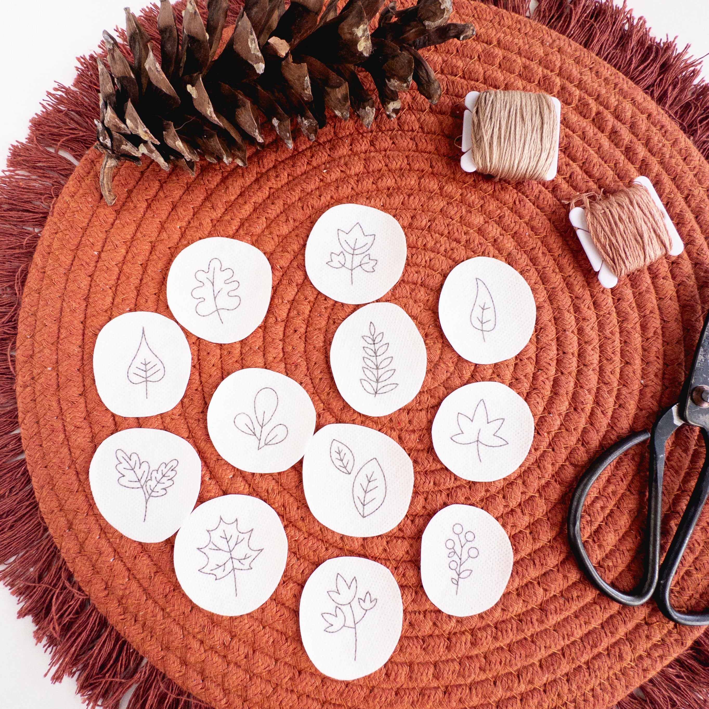 12 mini autumn botanical stick and stitch embroidery patterns on a rust-colored mat with a pinecone, thread, and scissors.