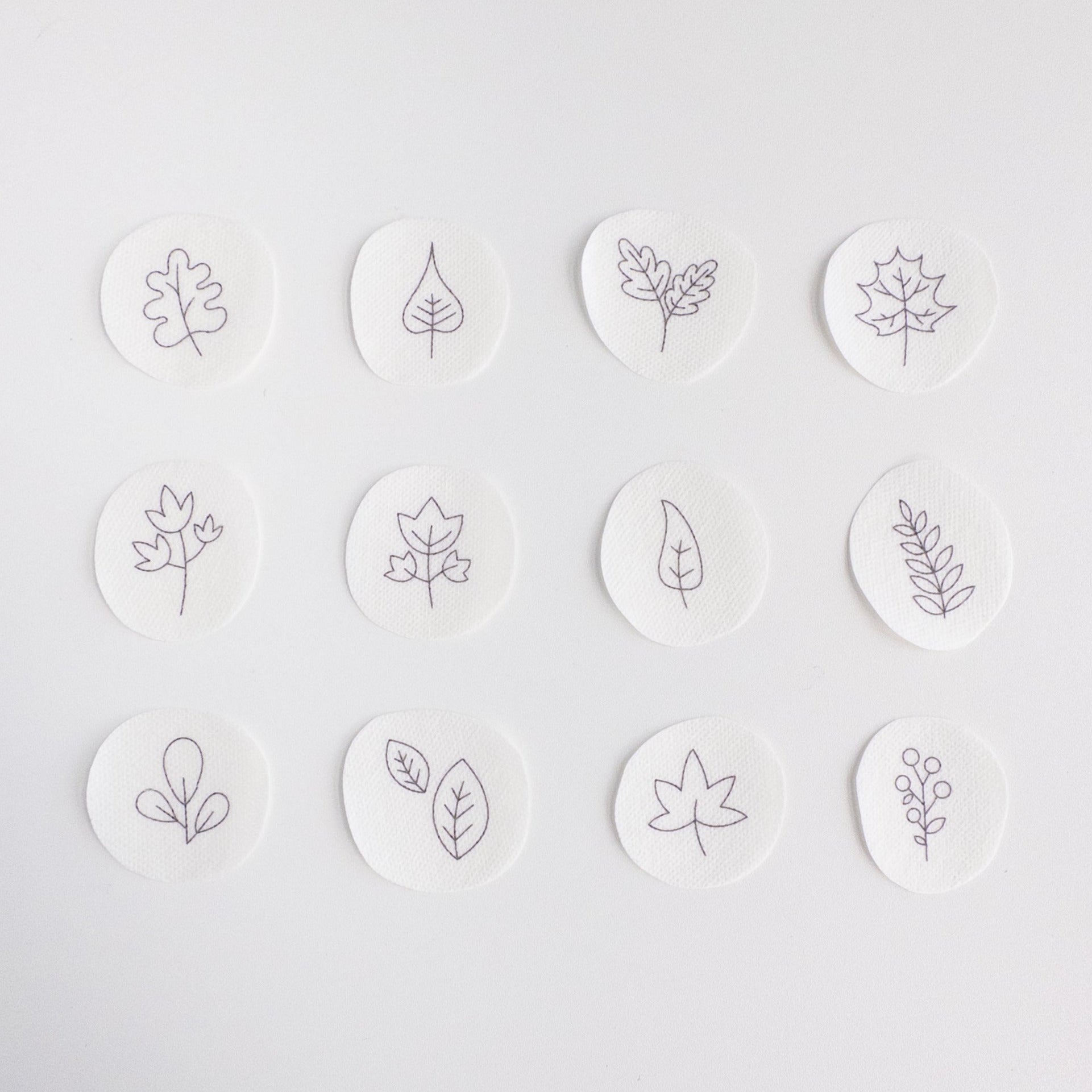 Twelve small, white, circular water-soluble patches with black outline autumn leaf and botanical embroidery patterns.