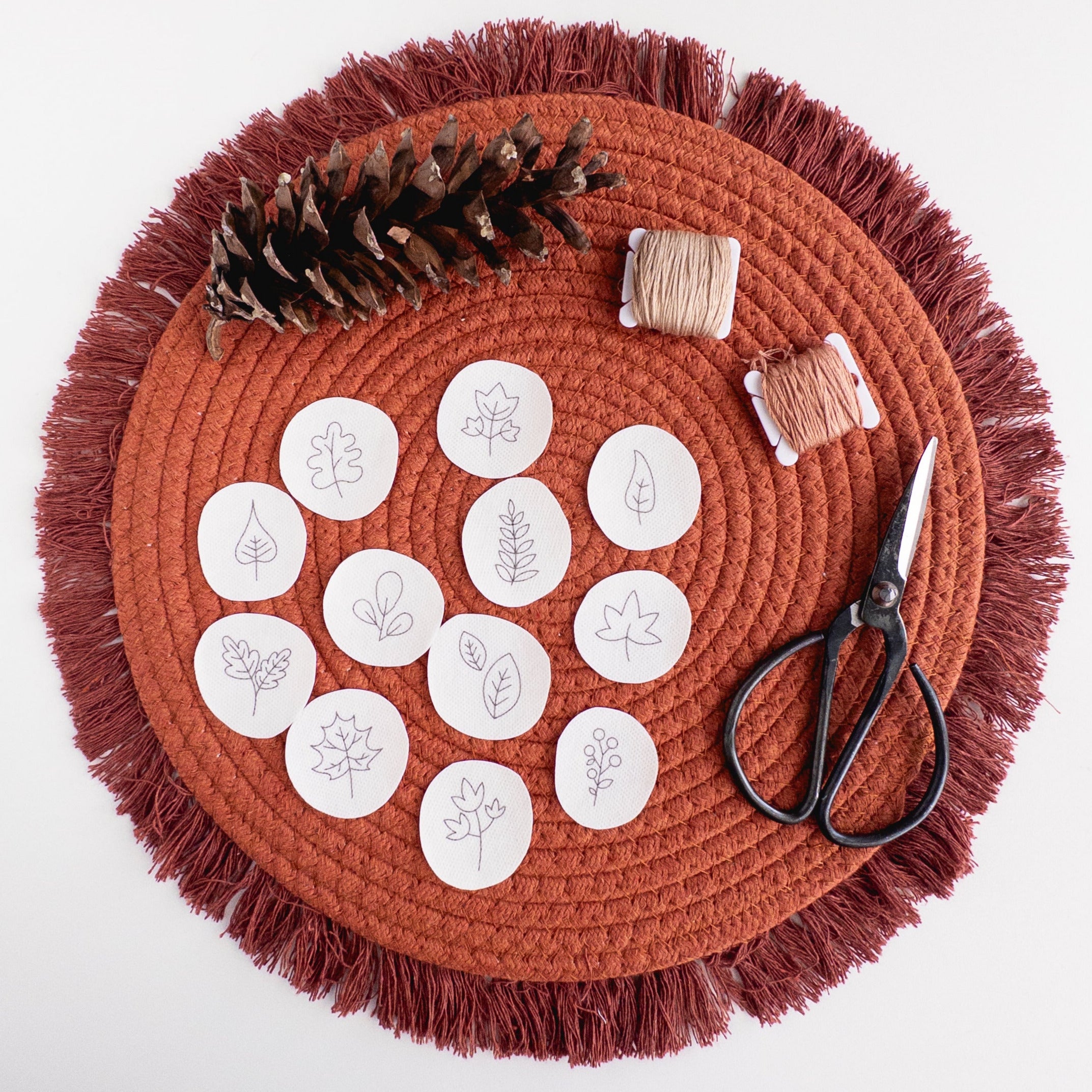12 mini autumn botanical stick and stitch embroidery patterns on white water-soluble stabilizer, with thread and scissors on a terracotta mat.