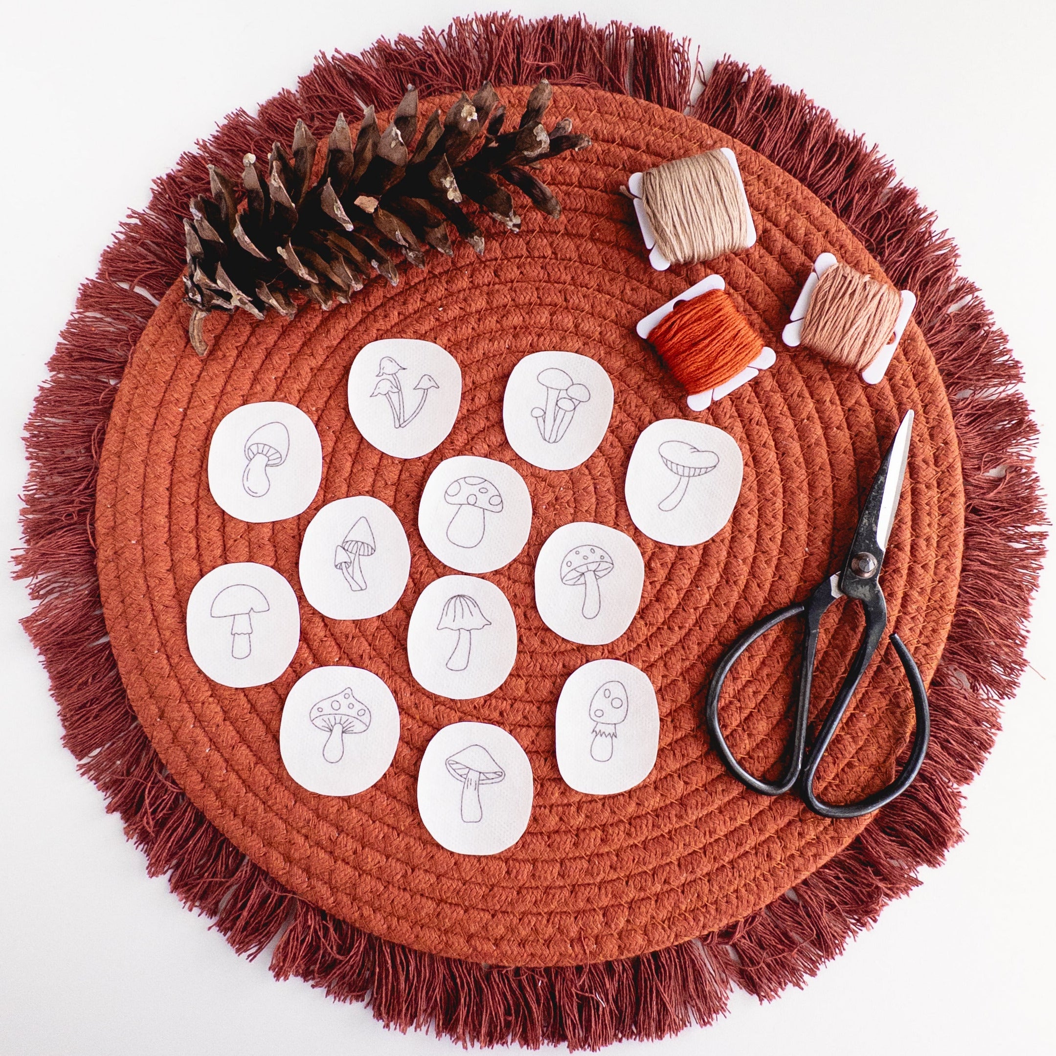 12 mini mushroom stick and stitch embroidery patterns on a terracotta mat with thread, pinecone, and scissors.