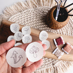 Hand holding two white water-soluble mushroom embroidery patterns, surrounded by more patterns, floss, and scissors.