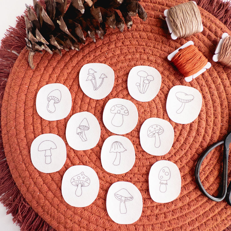 Twelve mini mushroom stick and stitch embroidery patterns on white water-soluble stabilizer, on a rust-colored mat.