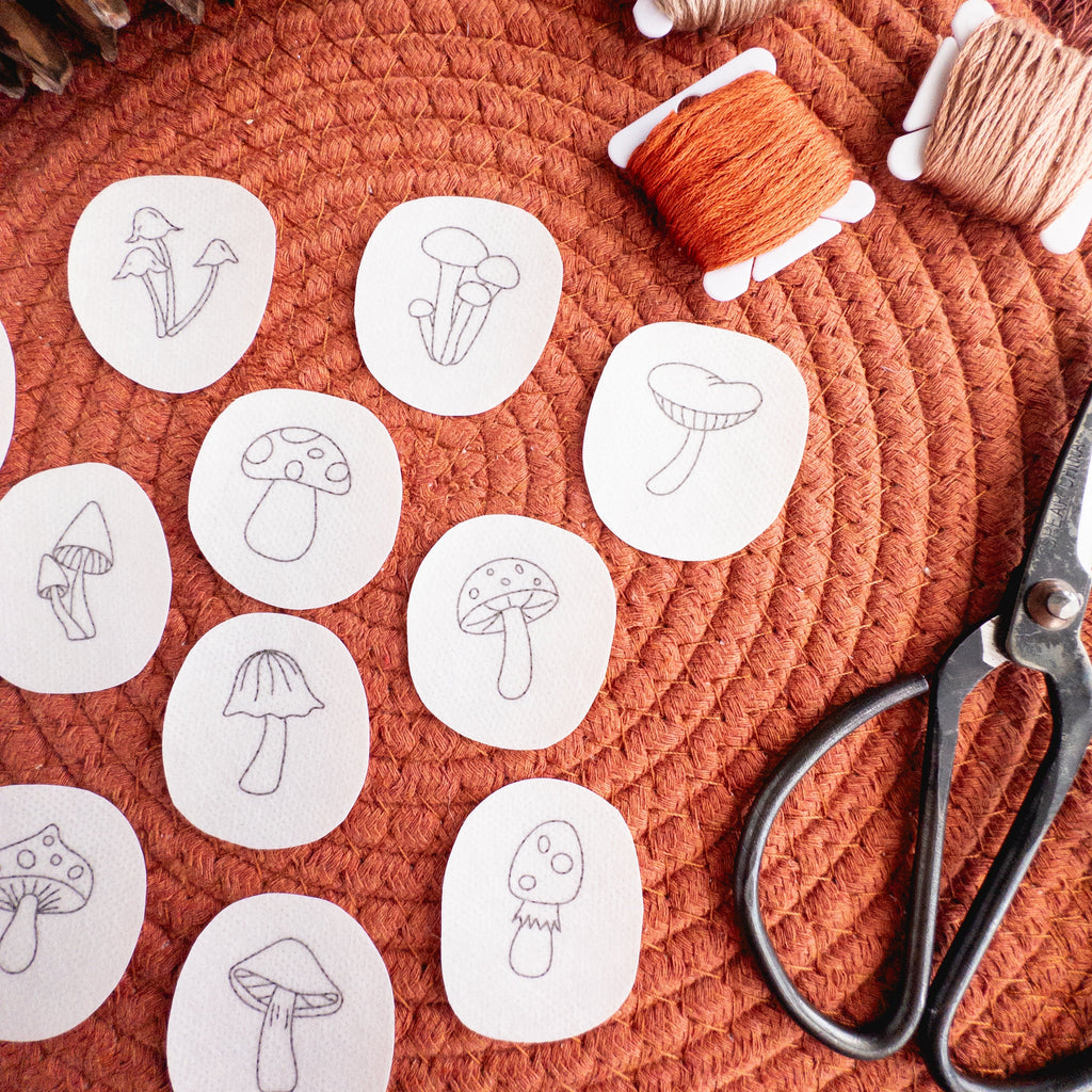 Twelve mini mushroom embroidery patterns on white water-soluble stabilizer patches, with orange and beige floss and scissors on a rust-colored mat.