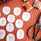 Twelve mini mushroom embroidery patterns on white water-soluble stabilizer patches, with orange and beige floss and scissors on a rust-colored mat.