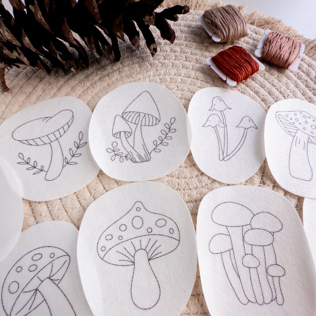 Nine white, water-soluble mushroom embroidery patterns on a woven mat with pinecones and brown floss.