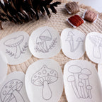 Nine white, water-soluble mushroom embroidery patterns on a woven mat with pinecones and brown floss.