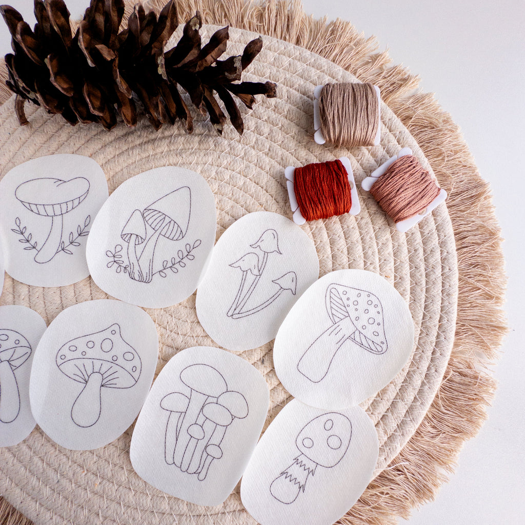 Nine mushroom stick and stitch embroidery patterns on white water-soluble stabilizer, with brown, red, and peach floss.