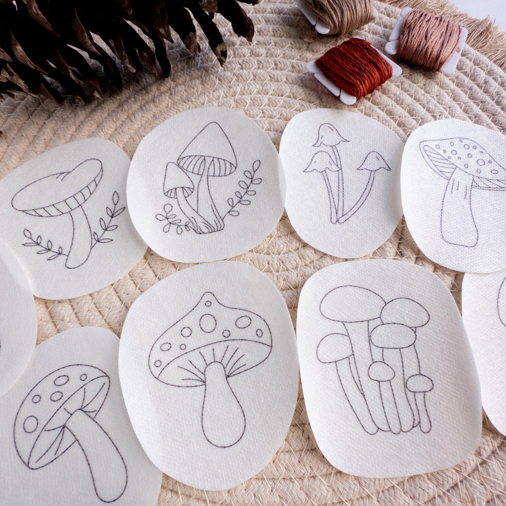 Nine mushroom stick and stitch embroidery patterns on white water-soluble stabilizer patches, with thread and pinecone.