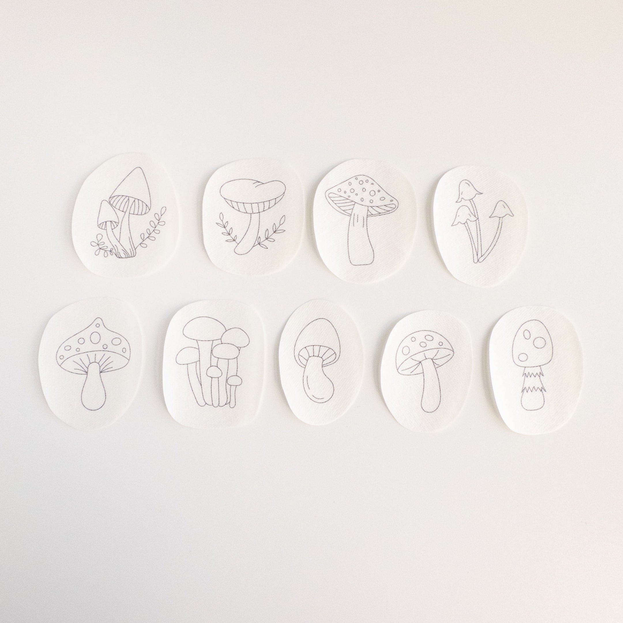 Nine oval, white water-soluble stabilizer patches with unique black line art mushroom embroidery patterns.