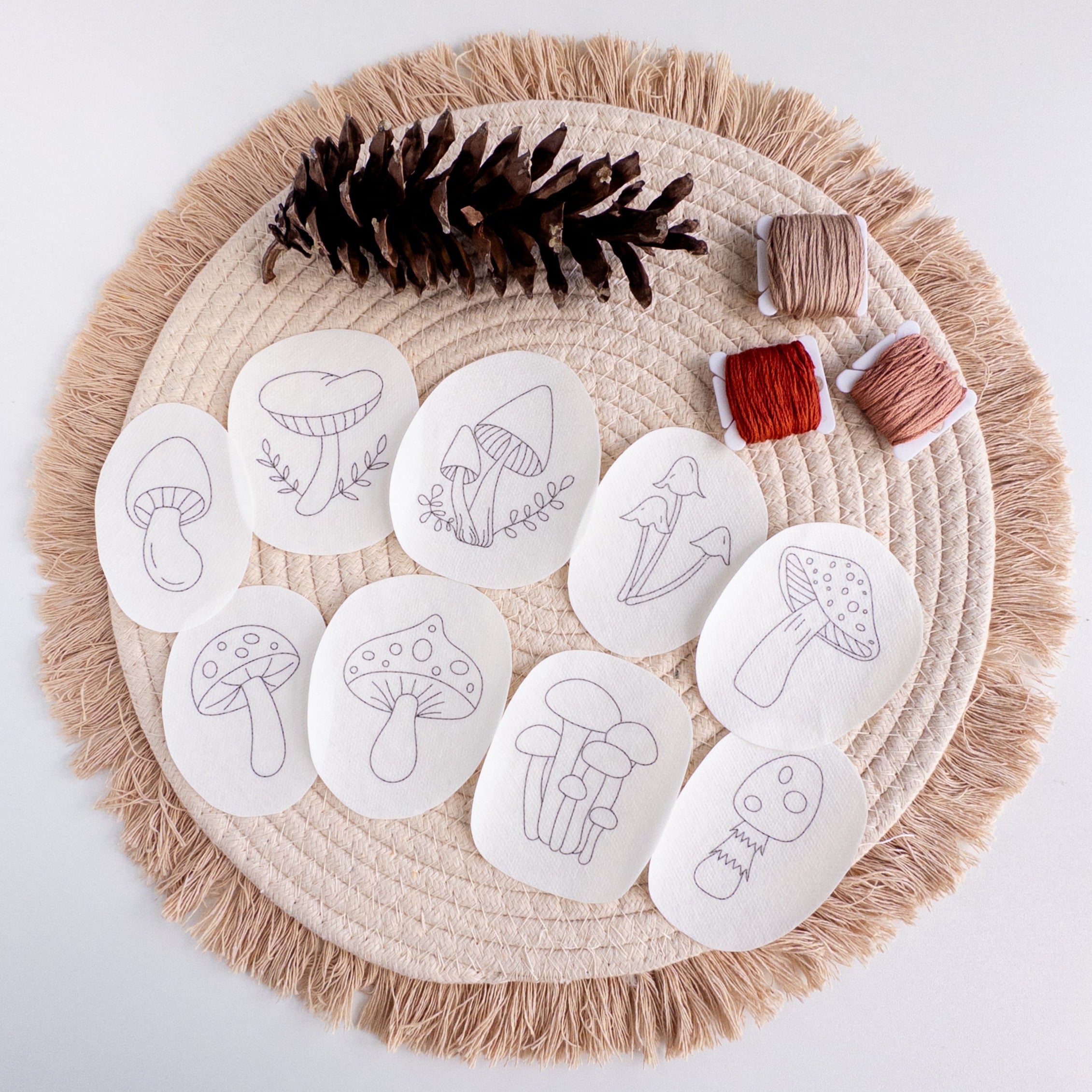 Nine mushroom stick and stitch embroidery patterns on white water-soluble stabilizer patches, with thread and pinecone.
