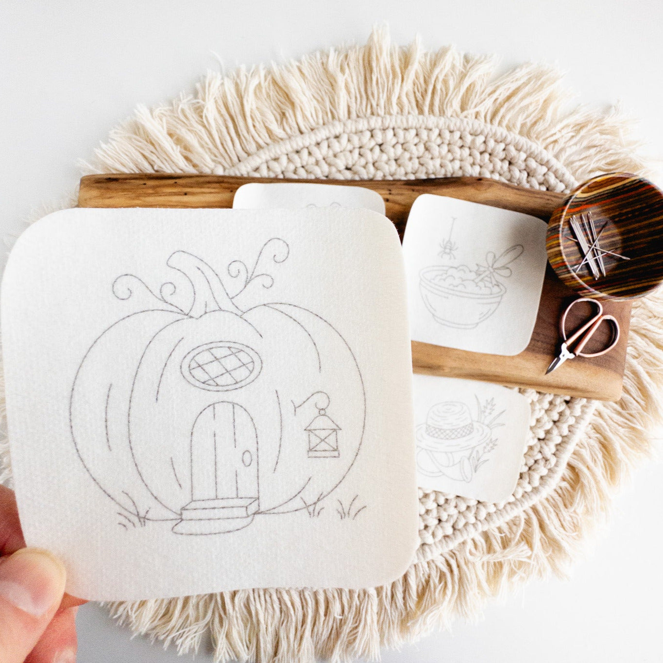 Hand holding a pumpkin house stick and stitch embroidery pattern, with other nursery rhyme designs, needles, and scissors.