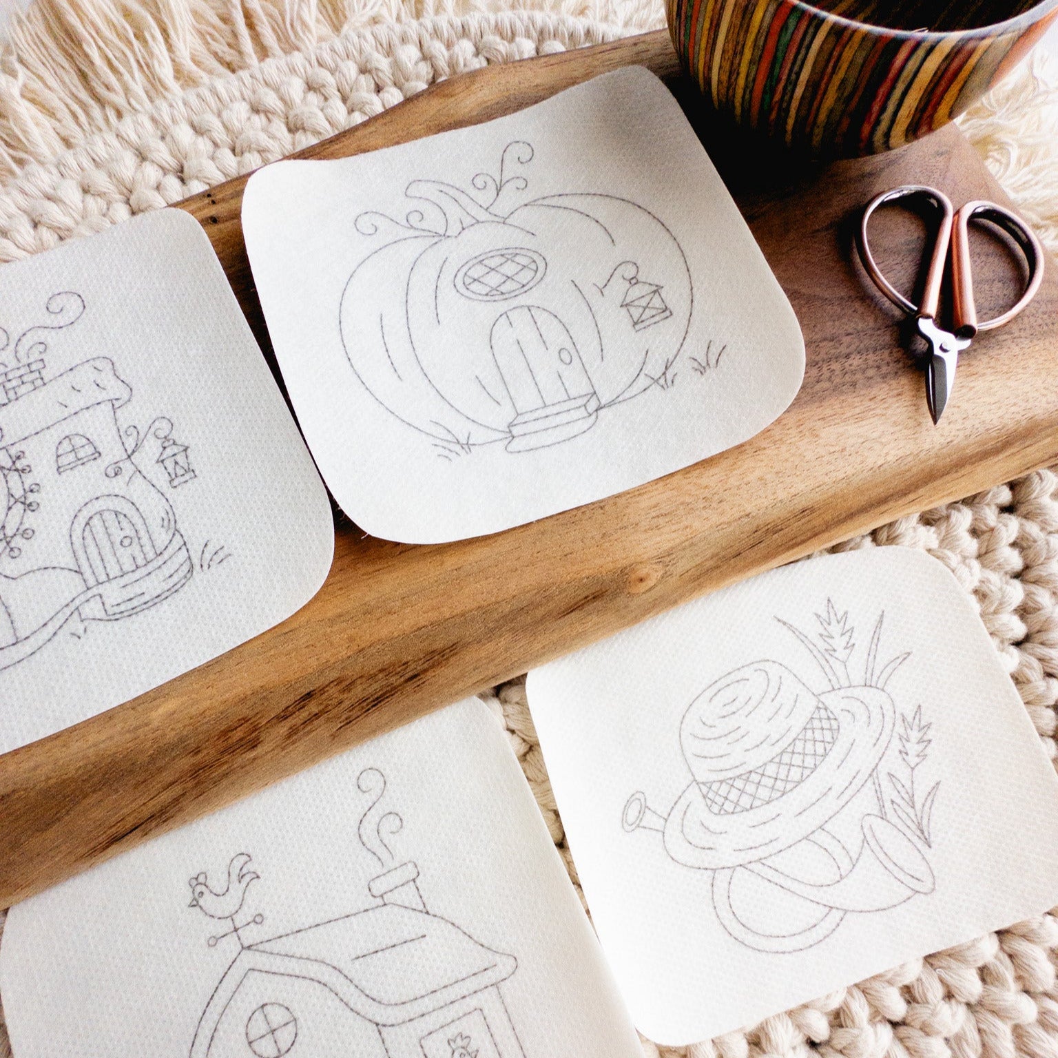 Four nursery rhyme stick and stitch embroidery patterns on water-soluble stabilizer, featuring whimsical designs like a pumpkin house and straw hat, on a wooden board with scissors.