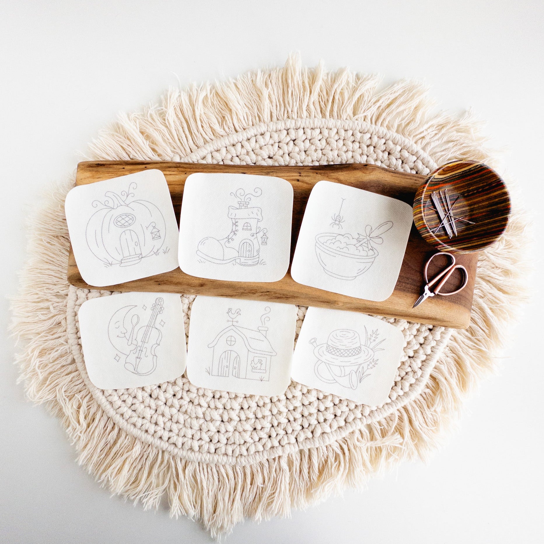 Six white stick and stitch nursery rhyme embroidery patterns on a wooden board with scissors and needles.