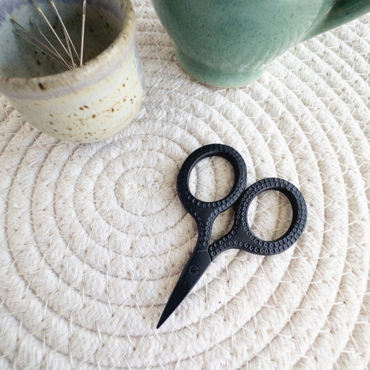 Black decorative scissors on a textured woven surface with a ceramic mug and container in the background.