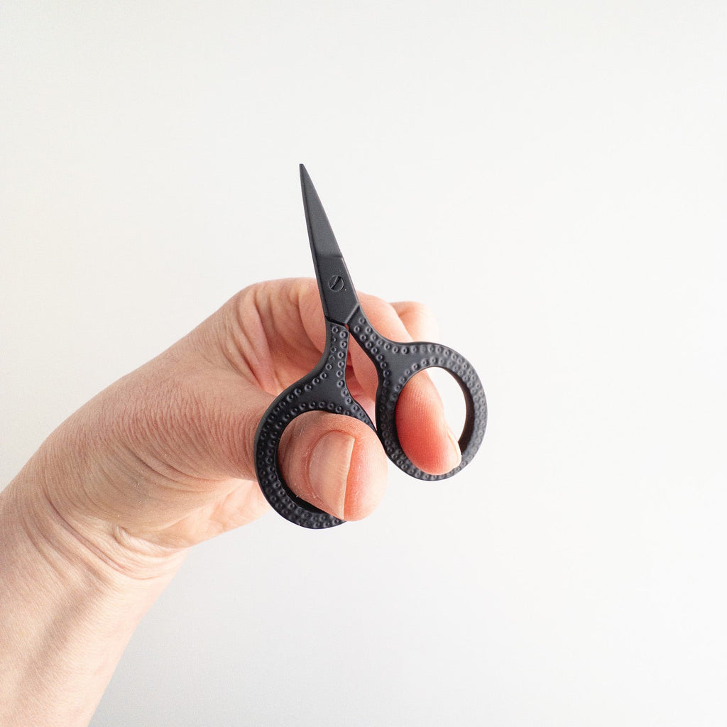 Hand holding a pair of black-handled, patterned scissors against a light background