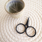 Black scissors and a ceramic bowl with embroidery needles on a textured white surface