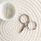 Small white bowl with embroidery needles and silver scissors on a textured beige surface