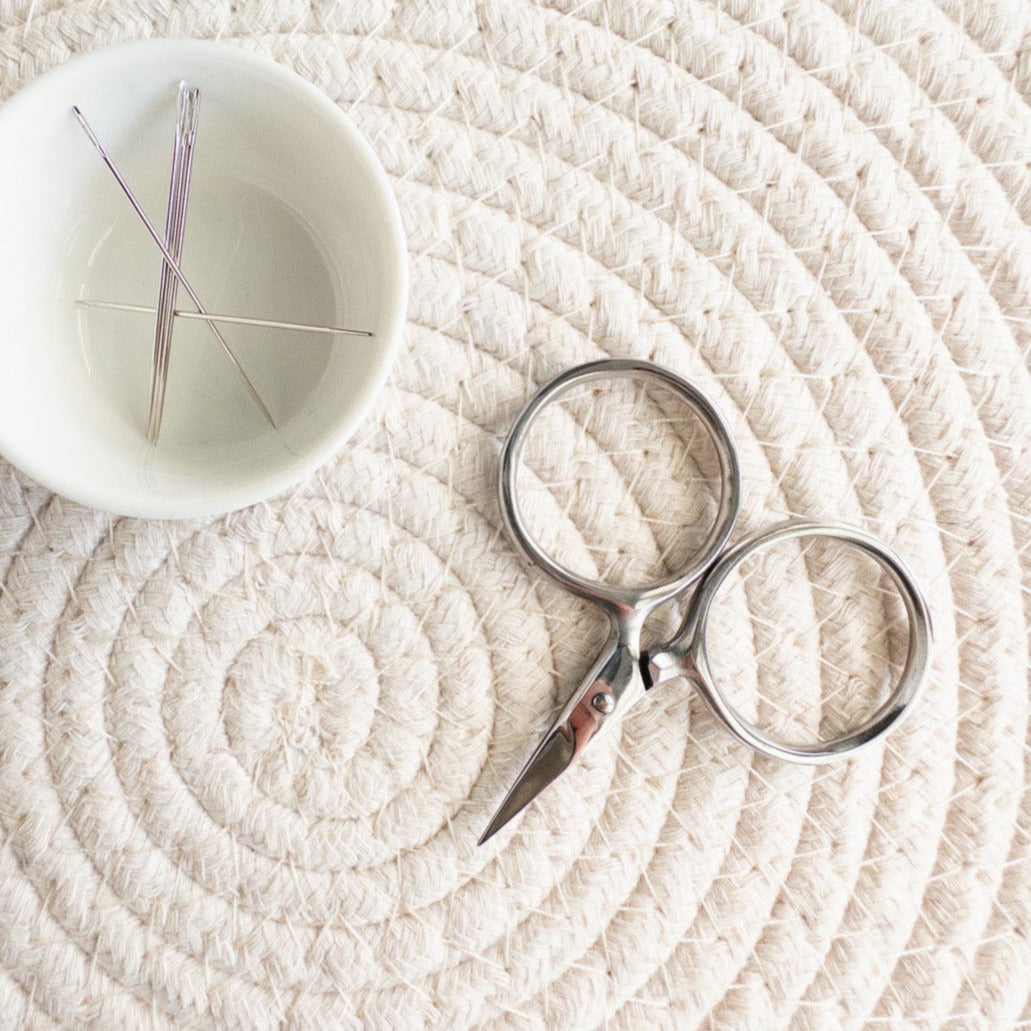 Small white bowl with embroidery needles and silver scissors on a textured beige surface