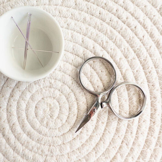 Small white bowl with embroidery needles and silver scissors on a textured beige surface