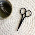 Black scissors on a textured white surface with a small bowl containing embroidery scissors.