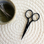 Black scissors on a textured white surface with a small bowl containing embroidery scissors.