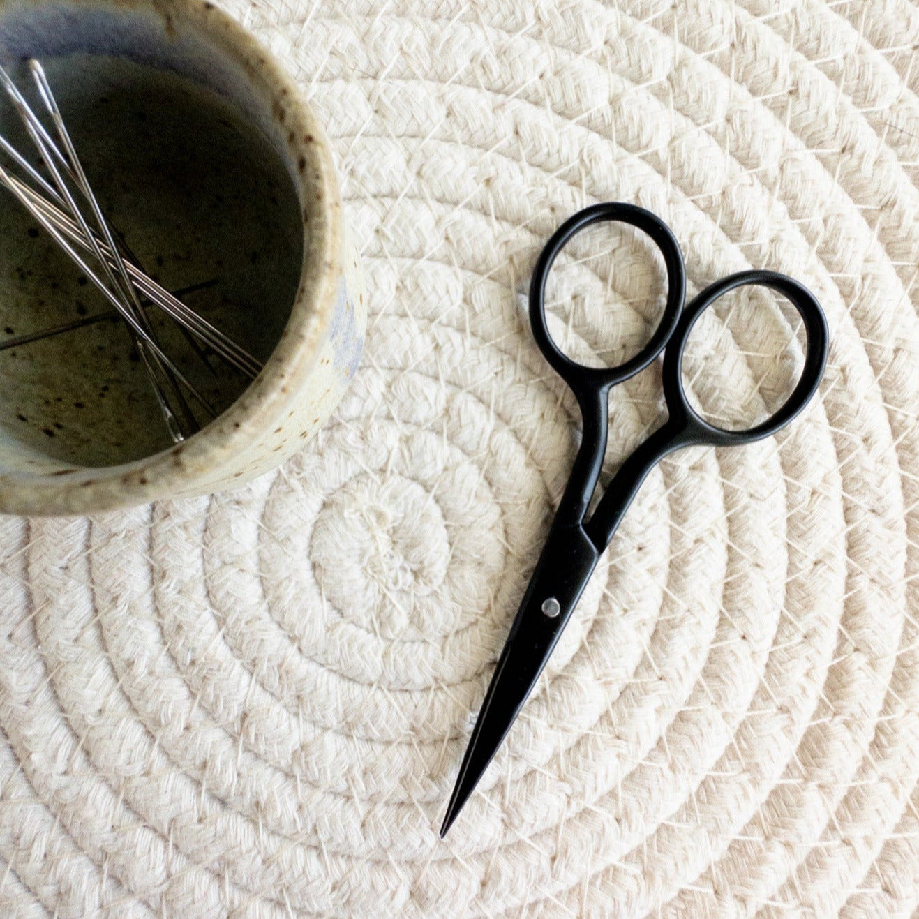 Black scissors on a textured white surface with a small bowl containing embroidery scissors.