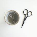 Small, round textured bowl with embroidery needles inside next to a pair of black scissors on a white background