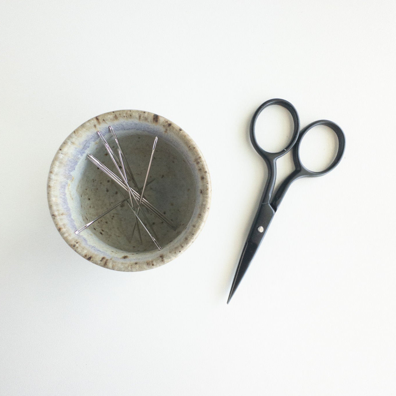 Small, round textured bowl with embroidery needles inside next to a pair of black scissors on a white background