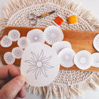 Hand holding a sunburst Stick and Stitch pattern, with more patterns, orange/yellow floss, and scissors on a wooden board.