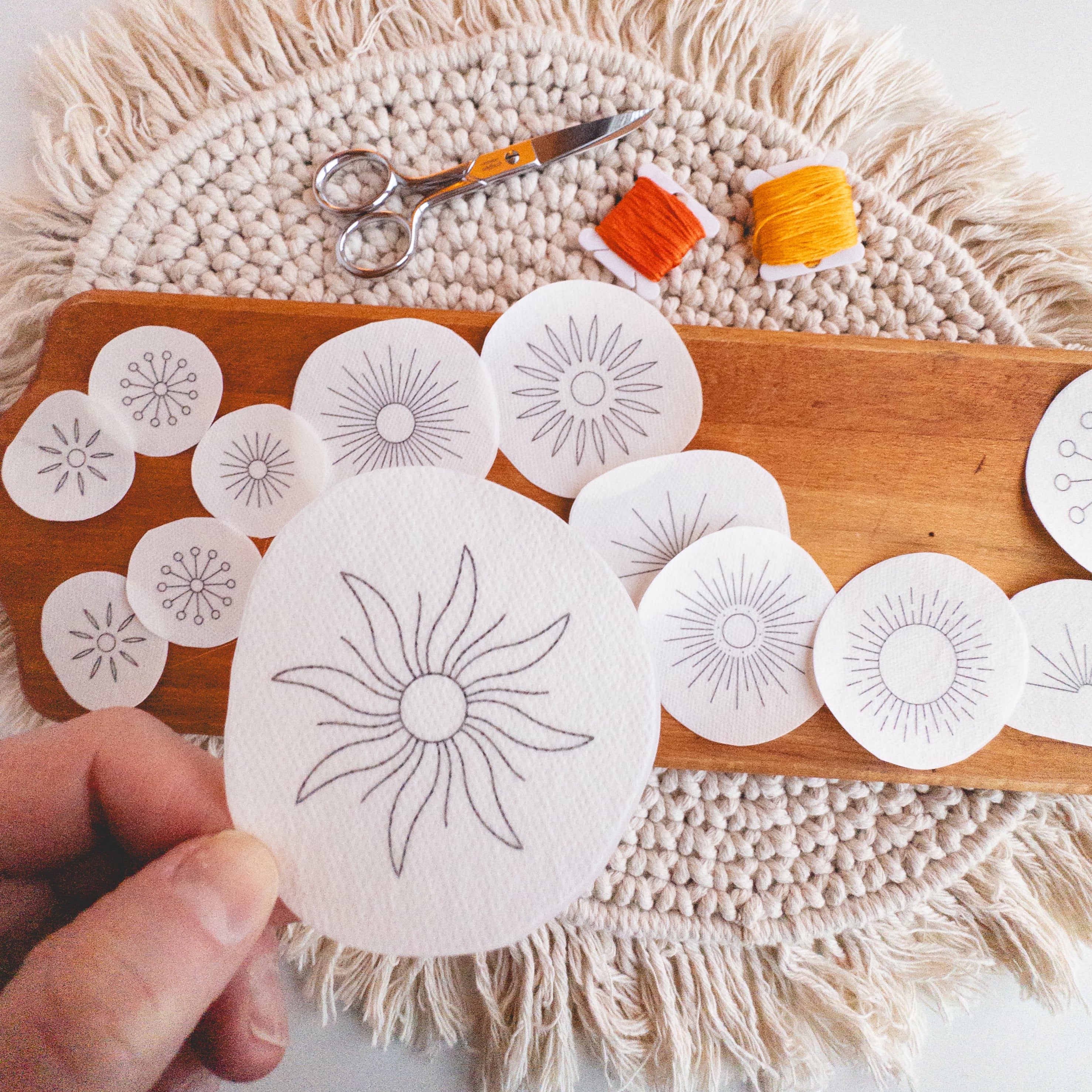Hand holding a sunburst Stick and Stitch pattern, with more patterns, orange/yellow floss, and scissors on a wooden board.